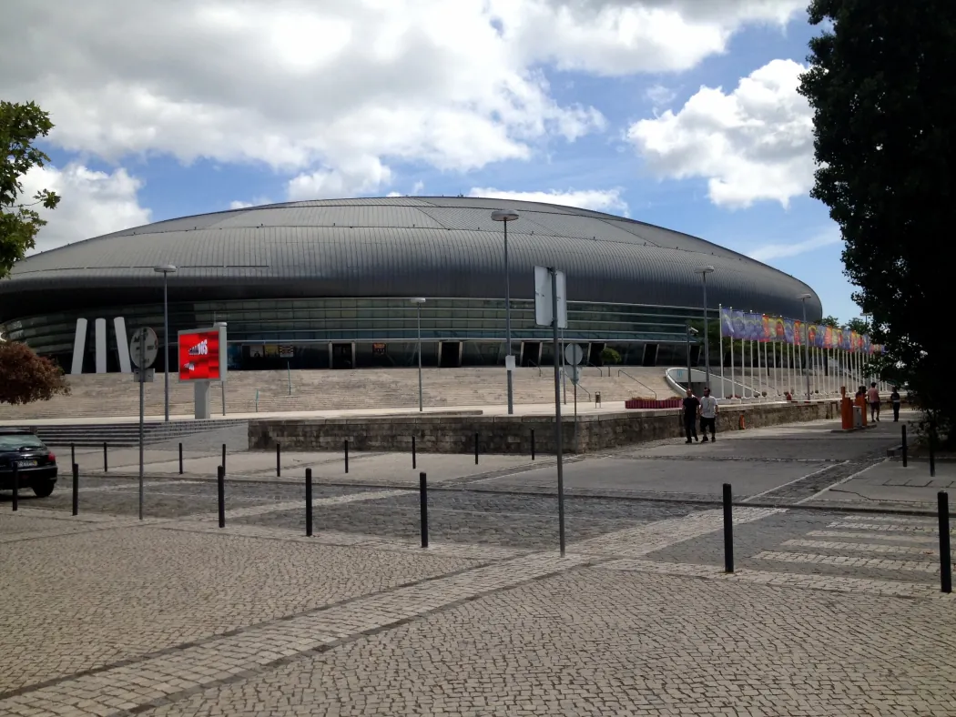 Exterior de uma grande arena coberta em Lisboa, com cobertura metálica curva, escadaria de acesso e fila de bandeiras