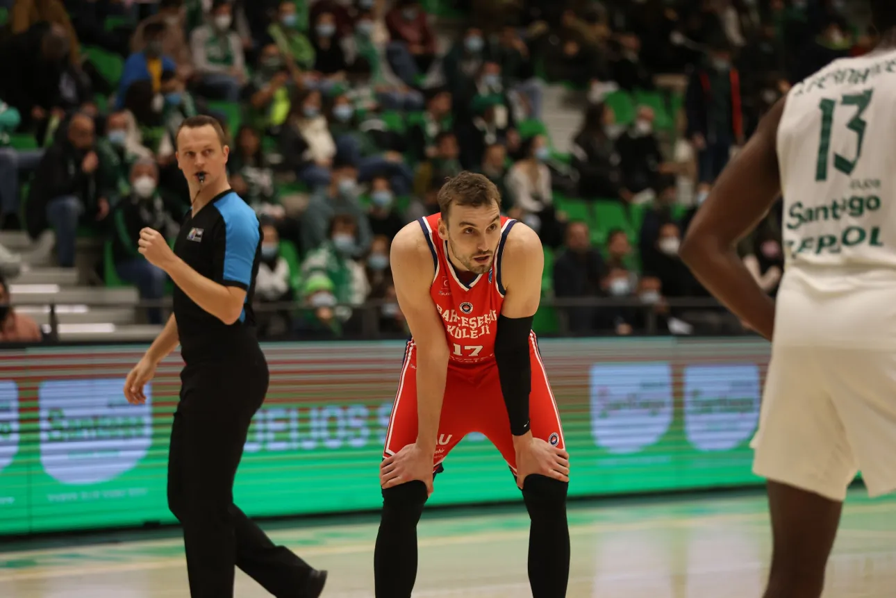 Pausa num jogo europeu de basquetebol: jogador de vermelho com as mãos nos joelhos, árbitro a caminhar e adeptos de máscara na bancada