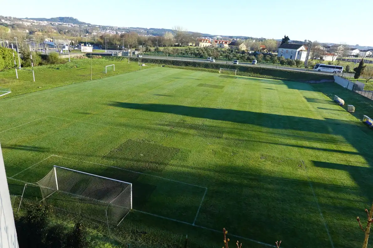 Campo de futebol de relva natural visto de cima, com a baliza e a grande área em primeiro plano e sombras longas da manhã