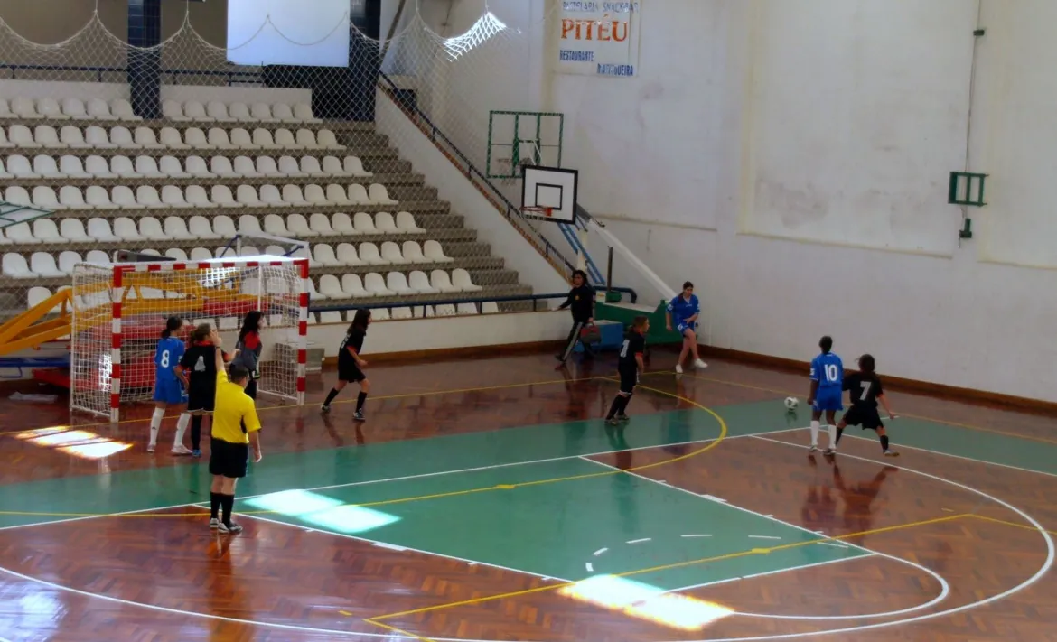 Jogo de futsal feminino num pavilhão de piso de madeira, equipa de azul contra equipa de preto junto à baliza, com o árbitro de amarelo