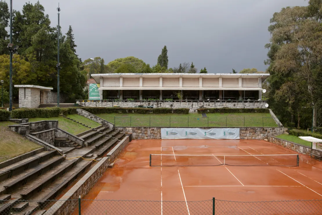 Court de terra batida molhado pela chuva com a rede montada, bancadas de pedra à esquerda e o edifício do clube sob céu carregado