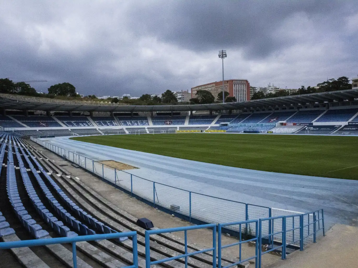 Bancada azul vazia do Estádio do Restelo com a pista de atletismo à volta do relvado, sob céu nublado