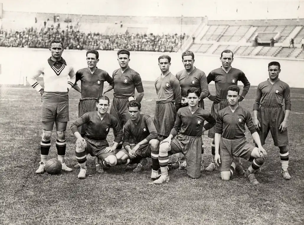 Fotografia a preto e branco da seleção portuguesa de futebol em 1928, com sete jogadores de pé e quatro de joelhos e uma bola de couro no relvado
